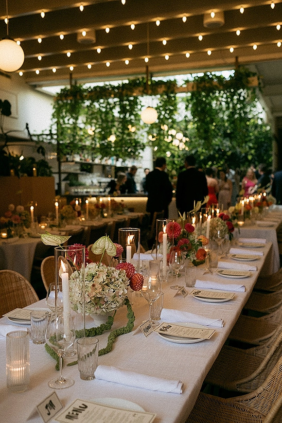 Reception tablescape on a long banquet table with colorful florals, taper candles, and greenery garland under string lights in an indoor space