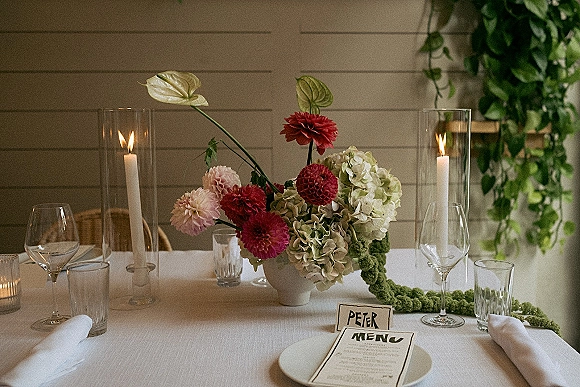 Reception tablescape with wedding table centerpiece of hydrangeas, dahlias, anthurium, taper candles in hurricanes, and menus on white linen