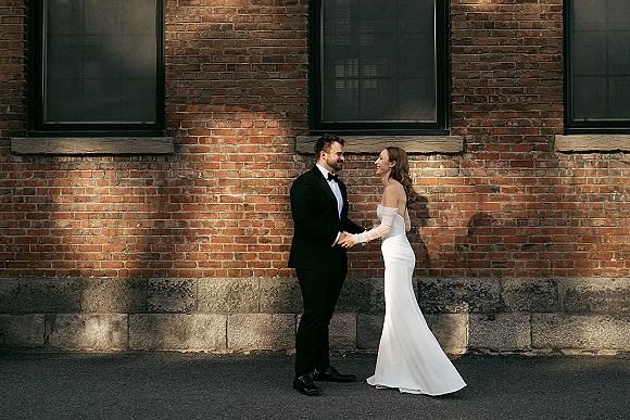 Couple portrait of bride and groom holding hands, laughing in front of a brick wall, her strapless dress with long gloves and his tuxedo bow tie