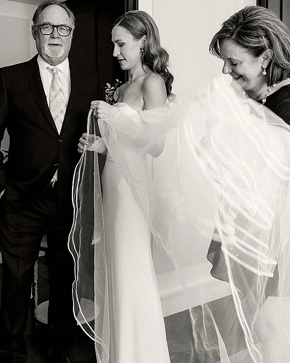 Bridal getting ready as the bride puts on veil while parents look on, wearing a strapless wedding dress in window-lit interior room