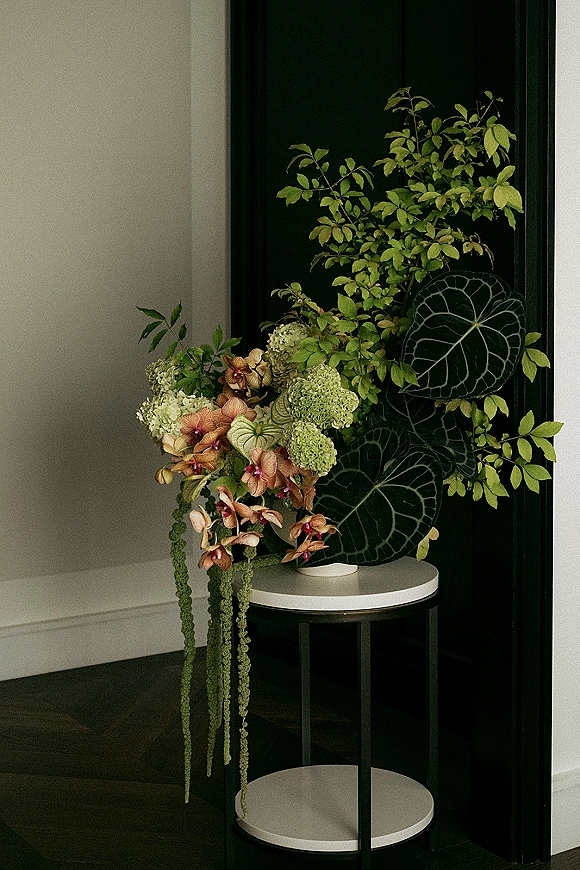 Wedding floral arrangement on a pedestal table with orchids, hydrangea blooms, and greenery against a dark paneled wall