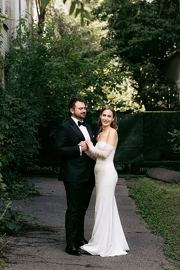 Couple portrait of bride and groom holding hands, her strapless dress with sheer sleeves beside his black tuxedo on a garden path