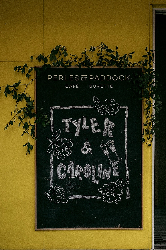 Wedding welcome sign on a chalkboard wedding sign with hand-lettered names, floral doodles, and greenery vines in a wood frame by a yellow doorway