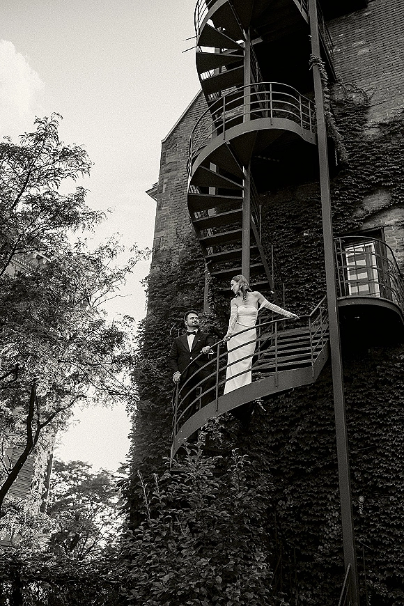 Couple portrait of bride in strapless wedding dress with long gloves and groom in tuxedo on an ivy-covered brick spiral staircase railing
