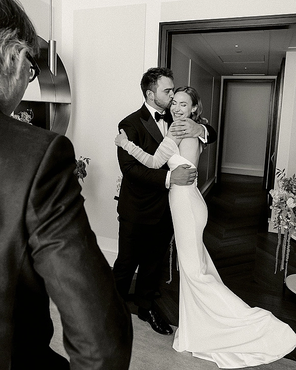 Wedding couple portrait of bride and groom hug as he kisses her forehead in a hotel hallway, her satin dress and opera gloves trailing behind