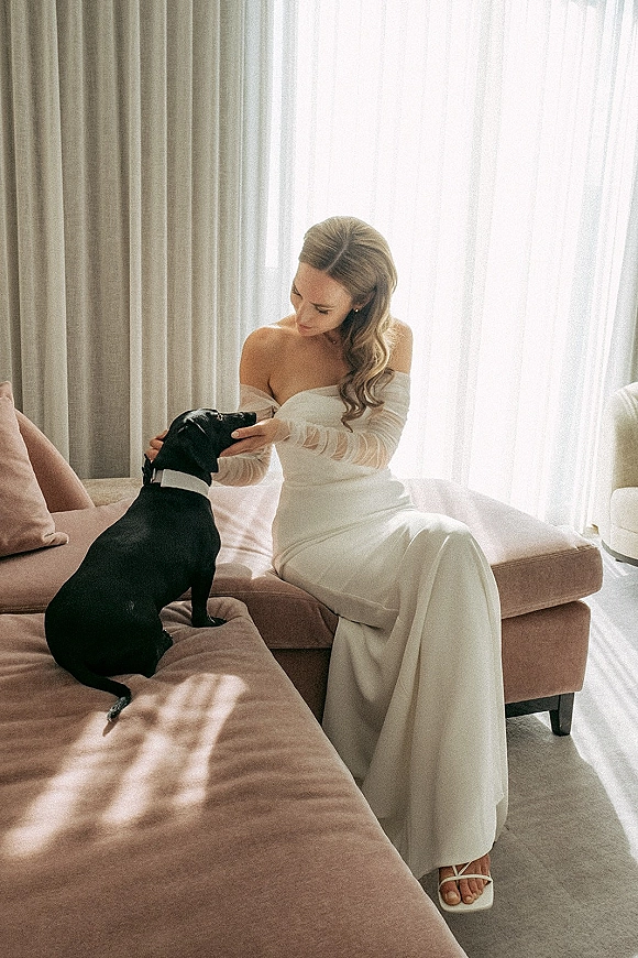 Bridal portrait of a bride with dog on a sofa, wearing an off-the-shoulder gown and long veil by sheer curtains in window light