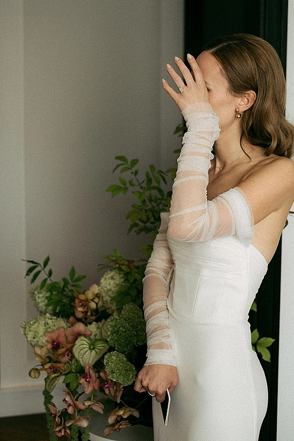 Bridal portrait of bride covering face pose, showing engagement ring close up in sheer opera gloves and off-shoulder dress by doorway