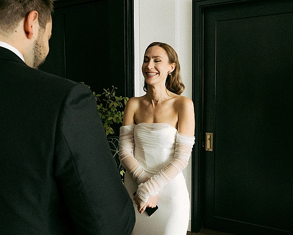 First look moment as the bride in a strapless dress and sheer opera gloves smiles at the groom by black paneled doorway, holding a phone