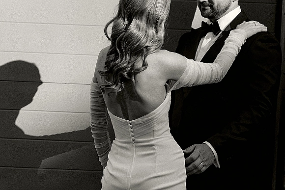 Couple portrait in a black and white wedding portrait style, bride in strapless button-back gown with tulle gloves embracing groom in tux by painted wood wall