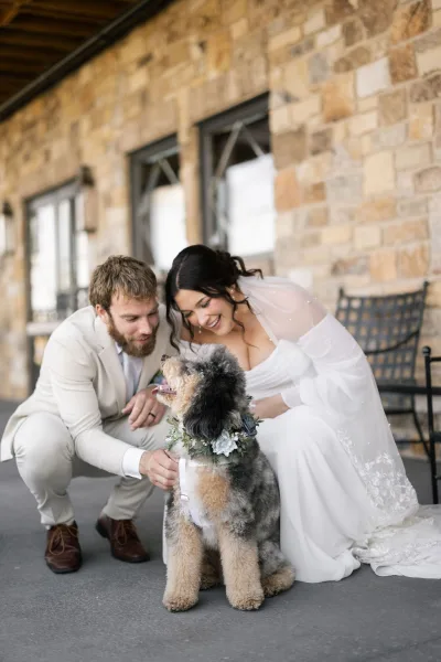 Couple with dog poses in a wedding dog photo as the bride pets their flower-collar pup beside a stone wall on a covered patio