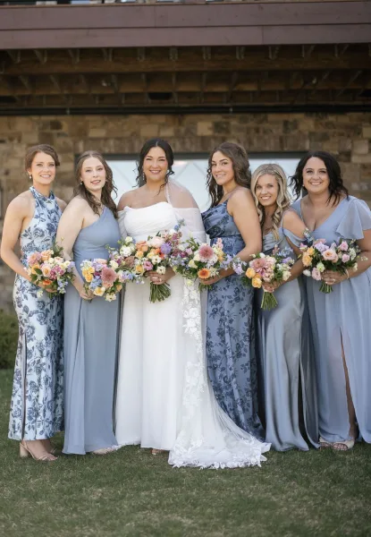 Bridesmaid group photo with bride in strapless gown and veil, bridesmaids in mismatched dresses holding bouquets by a stone wall pergola