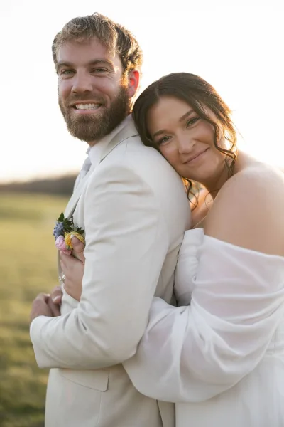 Couple portrait of bride hugging groom in a strapless gown as he smiles in a beige suit with boutonniere in an open field at golden hour
