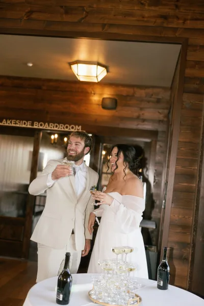Wedding toast as bride and groom clink champagne coupes beside a coupe tower on a white-clothed table in a wood-paneled indoor venue