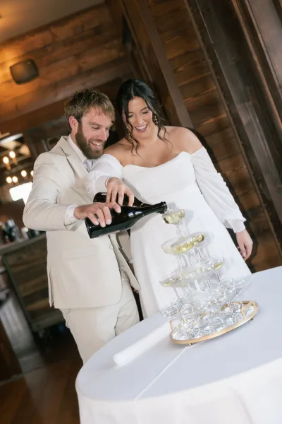 Champagne tower moment as bride in an off-shoulder dress and groom in a beige suit pour bubbles into coupe glasses indoors by a wooden wall