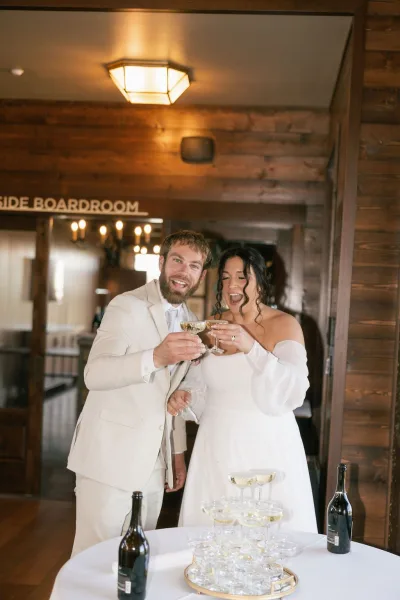 Wedding toast as bride in an off-the-shoulder dress and groom in a light suit raise champagne coupes beside a champagne tower in a wood-paneled reception