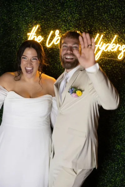 Couple portrait of bride and groom laughing and waving at a neon sign photo backdrop, groom showing wedding ring against a greenery wall