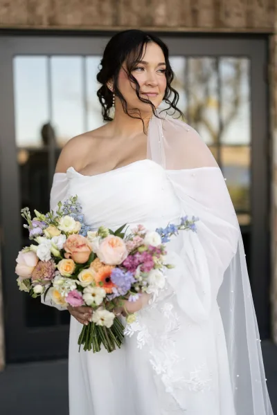 Bridal portrait of a bride in an off the shoulder wedding dress with lace applique, pearl-detailed tulle veil, holding a colorful bouquet by glass doors