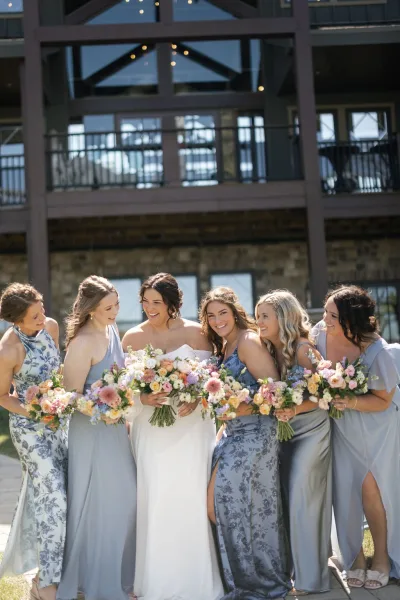 Bridesmaids group photo with bride holding bouquets in mix-and-match blue and gray dresses outside a modern lodge with string lights