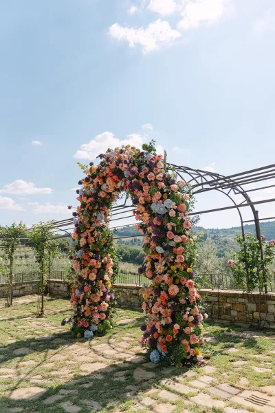 Floral wedding arch as an outdoor ceremony backdrop, lush with roses beneath a metal pergola, set against blue sky and distant hills