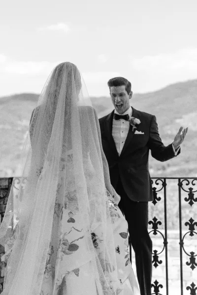 First look moment with a groom reaction photo as the bride in a floral bridal gown approaches on a mountain-view terrace with wrought iron railing.