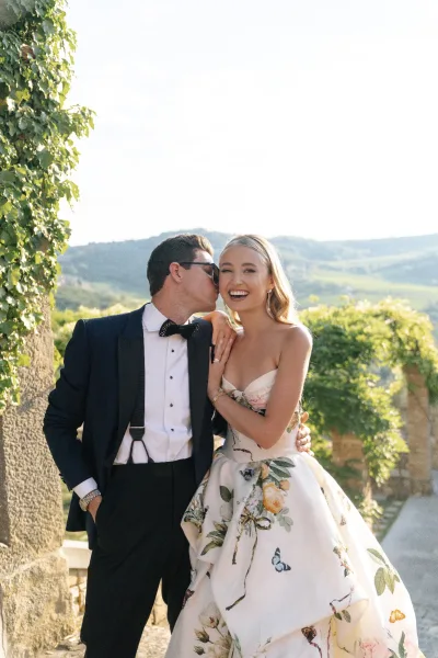 Couple portrait, groom kissing bride against an ivy-covered stone wall, his bow tie crisp as they smile amid climbing garden vines.