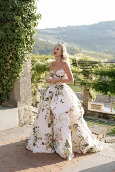 Bridal portrait of a bride in a floral wedding dress holding a champagne glass on a stone terrace beside an ivy-covered vineyard pergola.