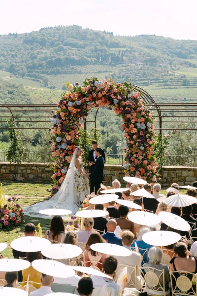 Ceremony moment during an outdoor wedding ceremony as bride and groom stand beneath a round floral arch, with vineyard hills beyond.