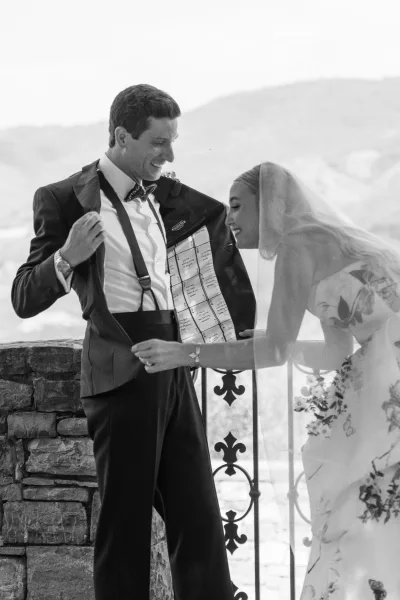 Candid wedding moment of the bride and groom laughing as he reveals his suit jacket lining on a stone terrace with a sweeping mountain view.