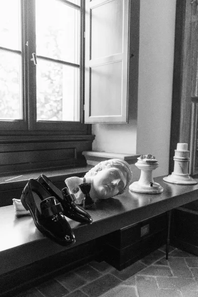 Groom details featuring black tie groom accessories—dress shoes, a classic bow tie, and a wristwatch set beside a statue bust.
