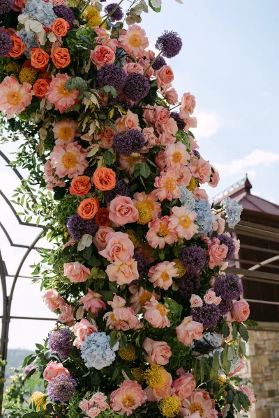 Floral wedding arch draped in lush ceremony arch flowers with roses, set against the sky, a stone building, and distant hills.