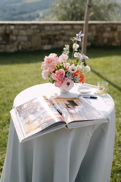 Wedding guest book displayed on a guest book table with a floral arrangement, set on a lawn before a stone wall and distant green hills.
