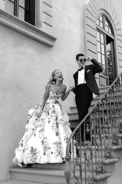 Couple portrait of a bride and groom walking on a stone staircase, groom with sunglasses, framed by a stucco wall and arched window.