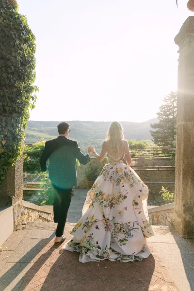 Floral wedding dress, a printed bridal gown, beside a groom in a suit on sunlit stone steps by an ivy wall with a hillside view.