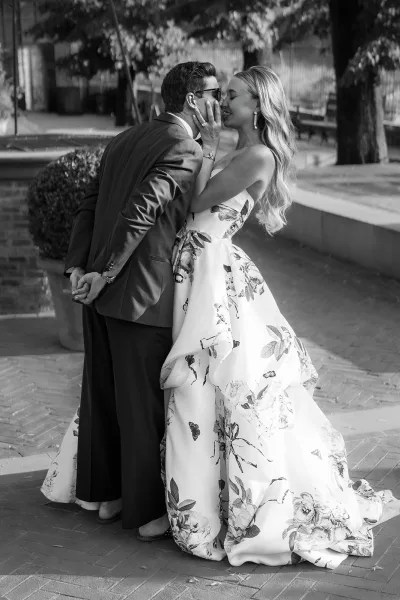 Black-and-white couple portrait capturing a candid couple moment as they lean close, groom in sunglasses, on a brick courtyard with potted topiary.