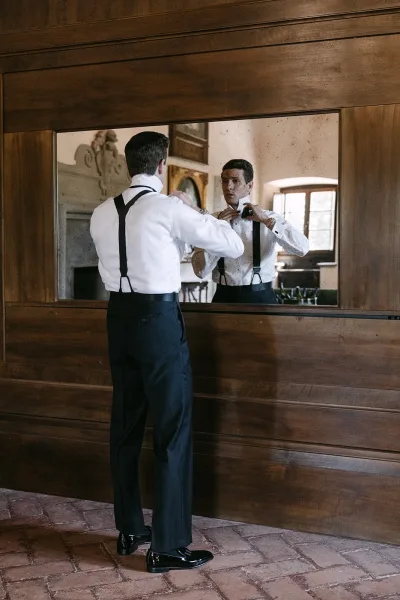 Groom getting ready, adjusting his bow tie in a wood-paneled room with brick floor and a fireplace in the background.