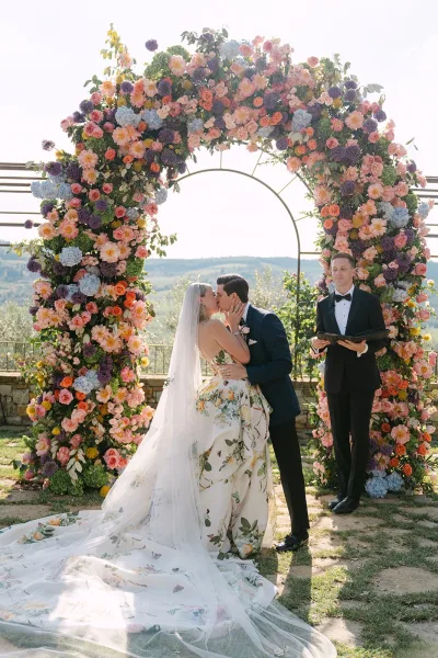Ceremony kiss moment as the couple shares their first kiss at the altar beneath a floral arch, with garden trees and hills in the background.