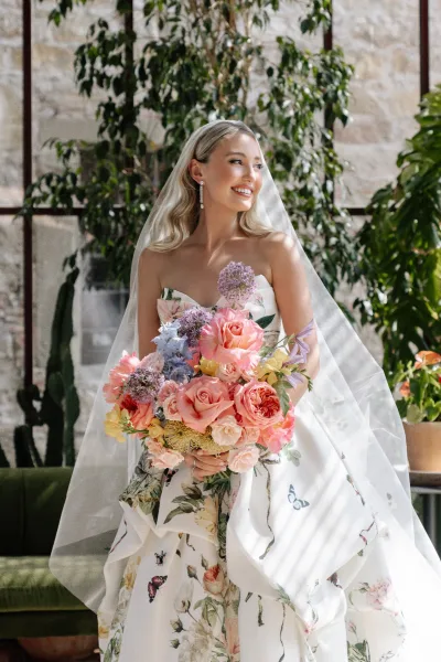 Bridal portrait of a bride with bouquet, wearing a strapless floral gown with butterfly print, posed in a sunlit indoor greenhouse.