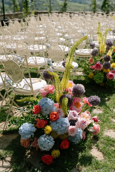 Ceremony aisle flowers frame outdoor ceremony decor, flanking wrought iron chairs beside a stone path with a hillside view.