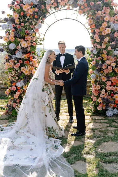 Ceremony moment with a couple exchanging vows beneath a floral arch, hands clasped, set against a hillside garden view and stone path.