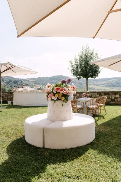 Wedding lounge seating for a garden cocktail hour centered on a tufted round banquette on a grass lawn against a low stone wall and hills.