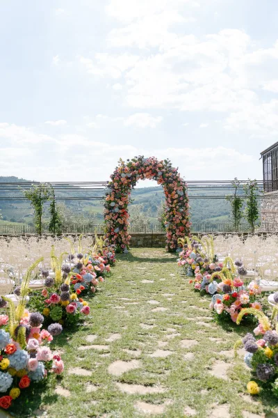 Floral wedding arch anchors an outdoor ceremony setup, framed by aisle flowers, with rolling hills and a stone wall beyond on the lawn.