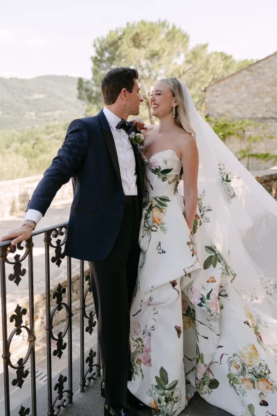 Couple portrait of bride and groom smiling, her long veil on a stone terrace with wrought iron railing and distant hills.
