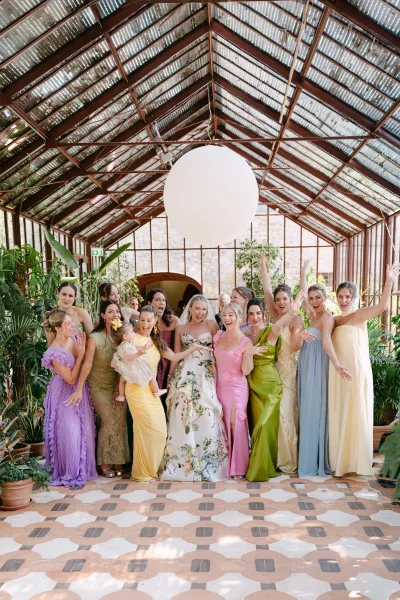 Bridal party portrait of bride with bridesmaids in mix-and-match pastel dresses, a flower girl’s bow, inside a sunlit greenhouse with glass walls.