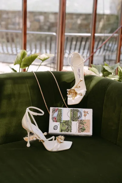 Bridal shoes, ivory bridal heels with floral appliqué, rest on a green velvet sofa beside indoor plants and a window.