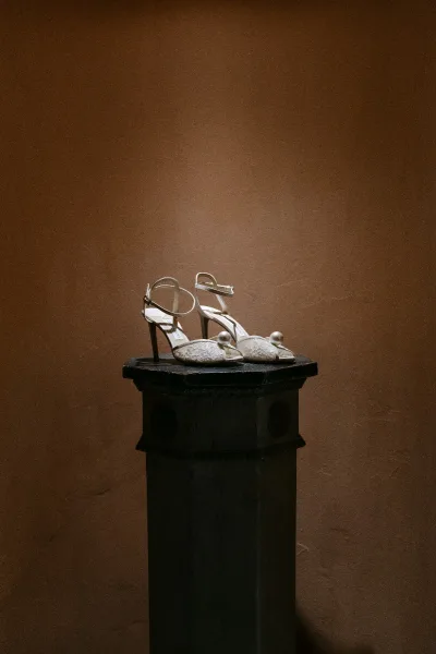 Bridal shoes, ivory wedding heels with pearl detailing, elegantly arranged against a brown plaster wall.