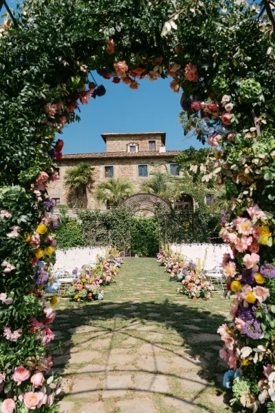 Ceremony setup with a floral arch frames a garden ceremony aisle before a stately stone building, bordered by garden trees under a blue sky.