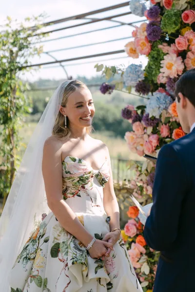 Wedding vows with the bride smiling at groom beneath a floral arch, set in garden greenery by a pergola with distant hills beyond.