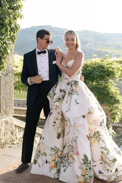 Couple portrait at an outdoor wedding, the bride in a floral gown, arm-in-arm on a stone terrace facing vineyard hills.