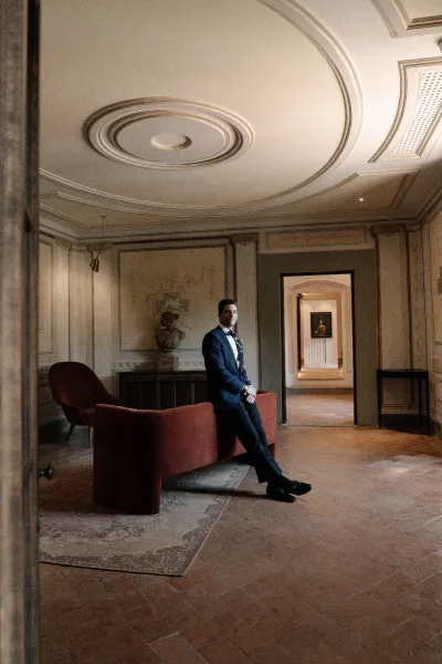 Groom portrait of a groom in tuxedo, seated on a velvet sofa in a historic interior with ornate ceiling molding and an arched doorway.