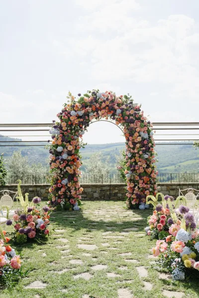 Floral wedding arch anchors an outdoor ceremony setup, framed by aisle flowers, with rolling hills and open sky beyond a low stone wall.
